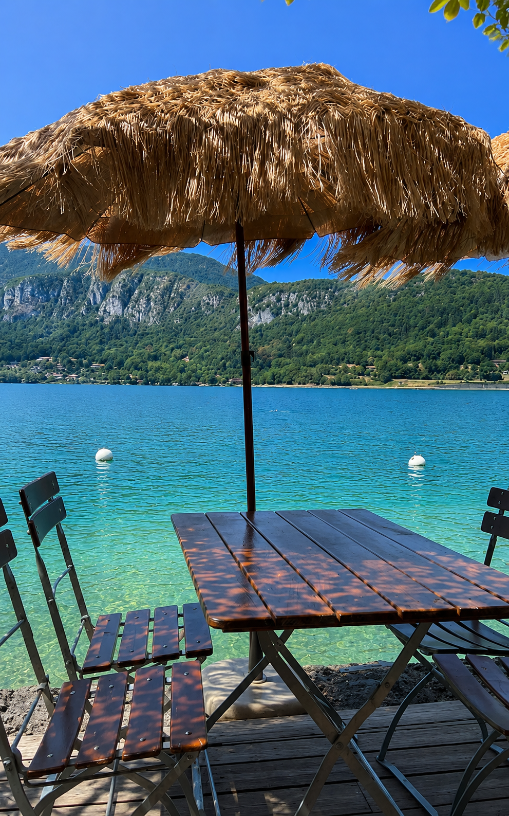 Table en bois sous parasol de paille face aux eaux turquoise du lac d'Annecy à Angon
